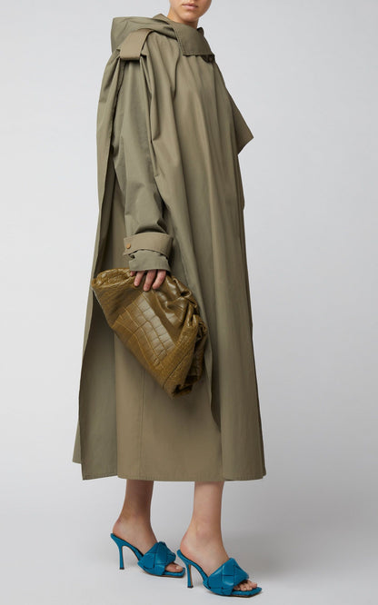 Person wearing a long olive green coat with Bottega Veneta Lido Mule Vintage Blue 608854VBSS04335 holding a textured bag on a white background