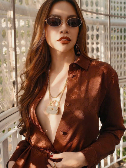 Hồ Ngọc Hà wearing a brown patterned shirt with Bottega Veneta Sardine BV1386S-001 Grey Gold Sunglasses 821495V44508043 and a necklace, standing outdoors.