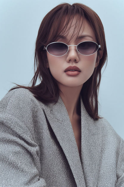 Woman wearing Bottega Veneta Sardine Oval Sunglasses - BV1386S 003 and a gray coat against a light blue background