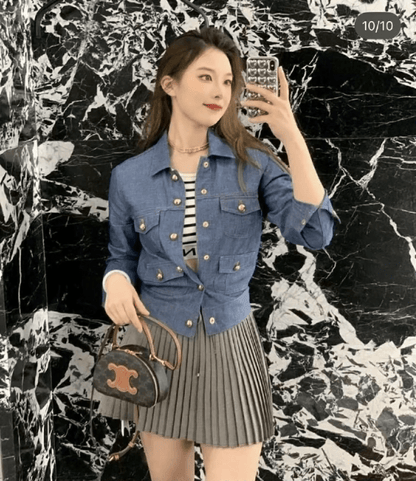 Woman taking a selfie wearing a blue denim jacket and pleated skirt and holding a Celine Half Moon Case In Triomphe Canvas & Calfskin 10K642ECM.04LU against a marble wall.