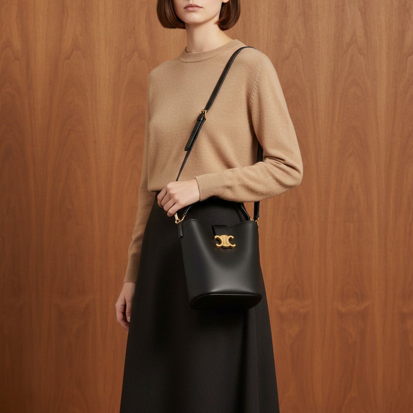 Person wearing a beige sweater and black skirt holding a Celine Small Bucket Louise In Smooth Calfskin Black 119173FX9.38NO with a gold clasp against a wooden background