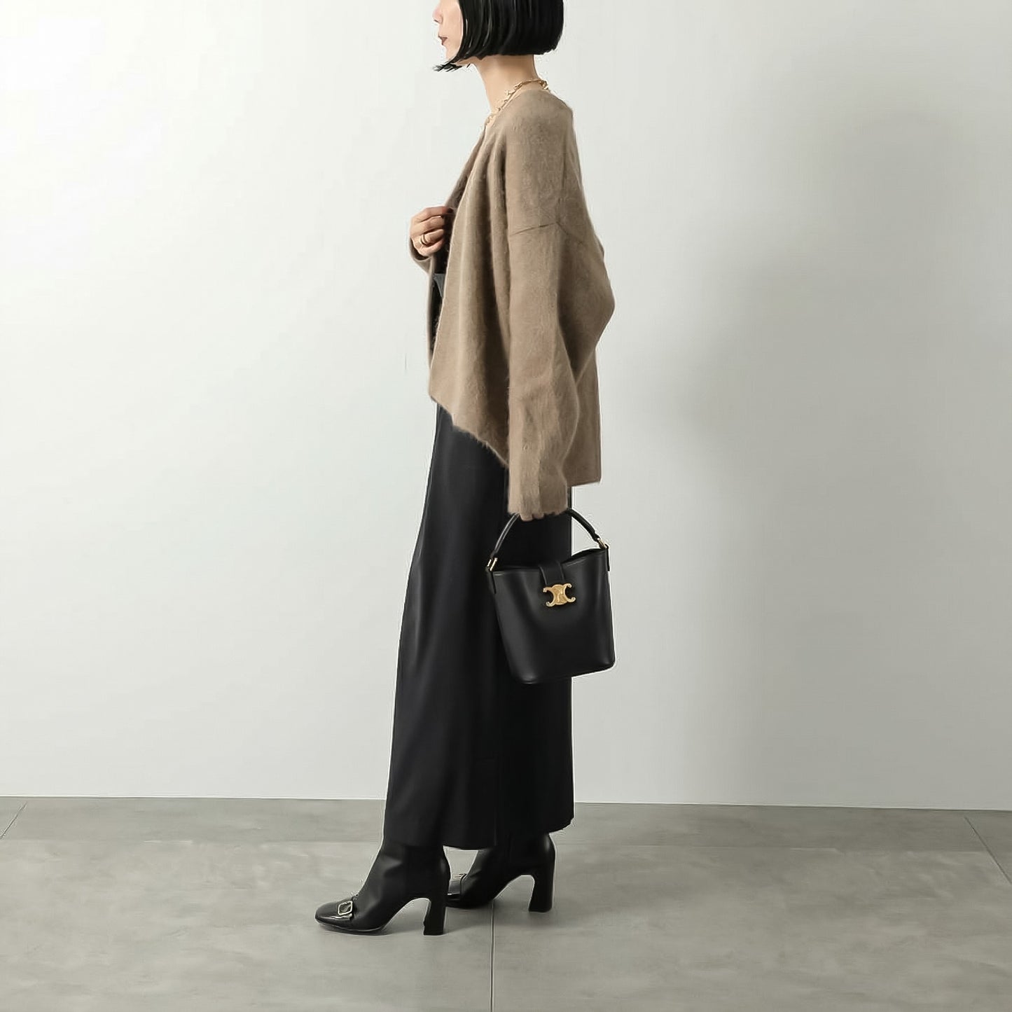Person wearing a beige cardigan and black pants holding a Celine Small Bucket Louise In Smooth Calfskin Black 119173FX9.38NO against a plain background