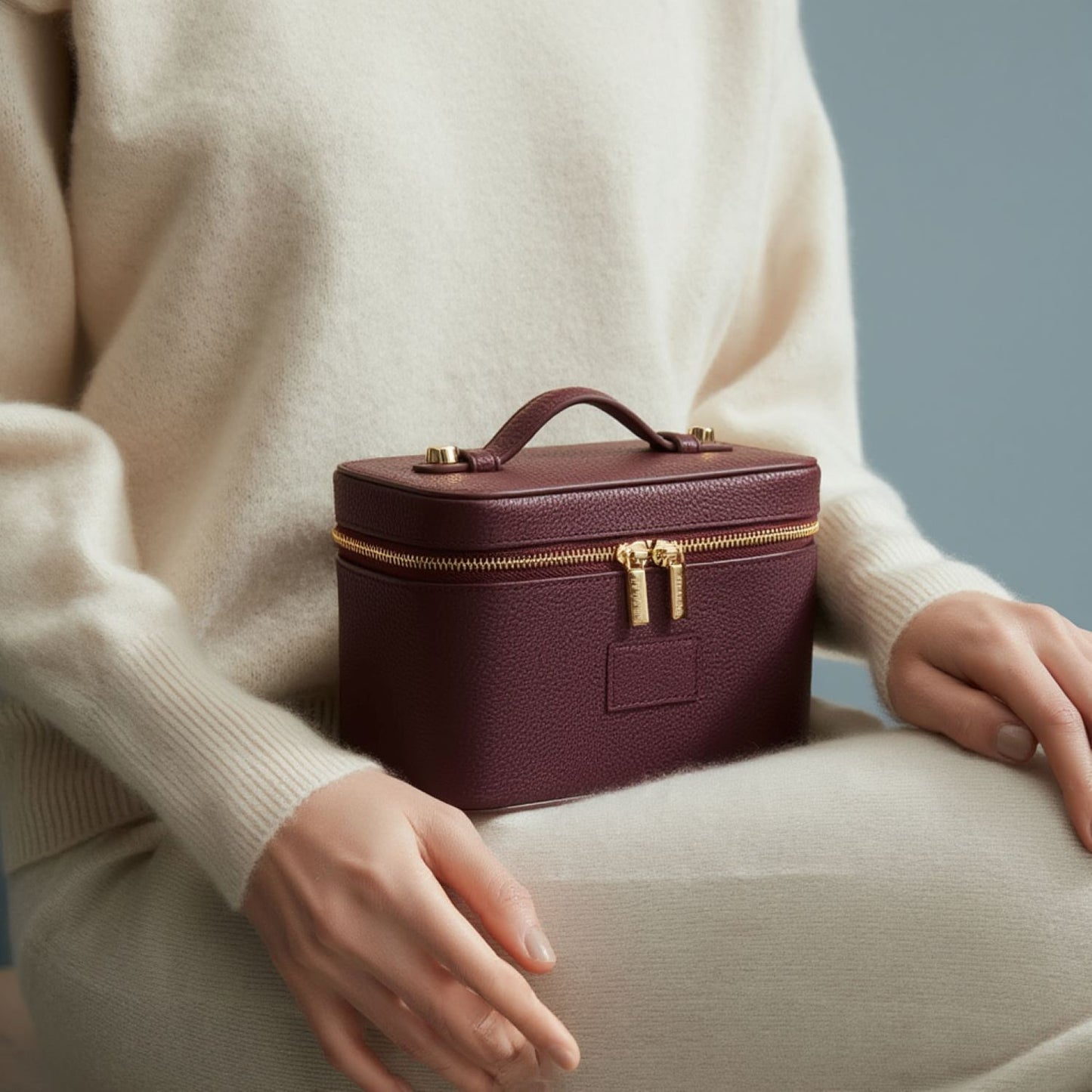 Etoile Mini Vanity Case Burgundy with gold zippers held by a person wearing a beige sweater.
