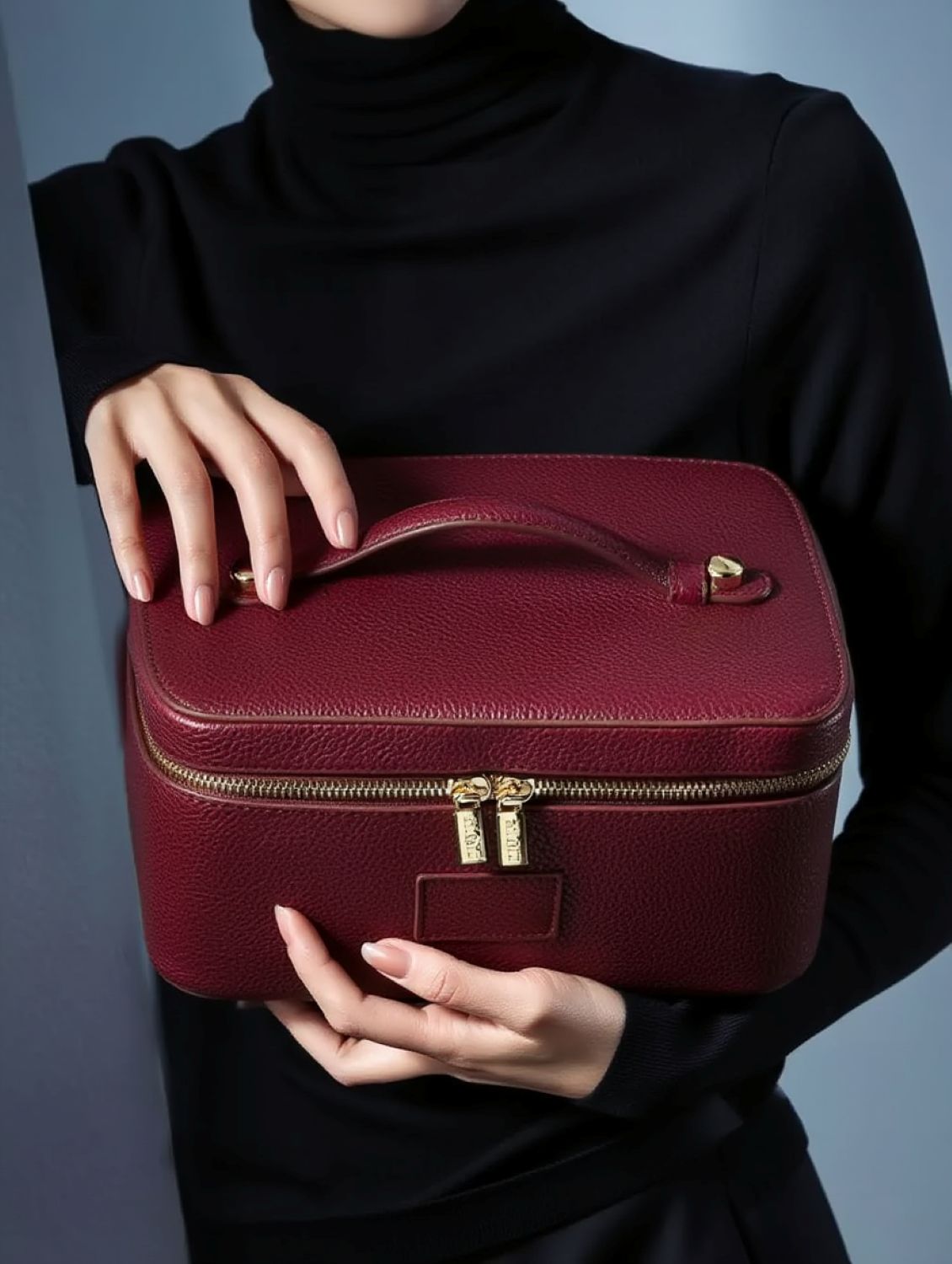 Person holding a Etoile Vanity Case Burgundy with gold zipper and clasp against a neutral background