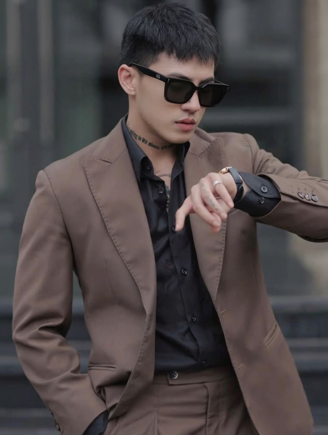 Man wearing a brown coat and Gentle Monster Daze 01, looking at his watch.