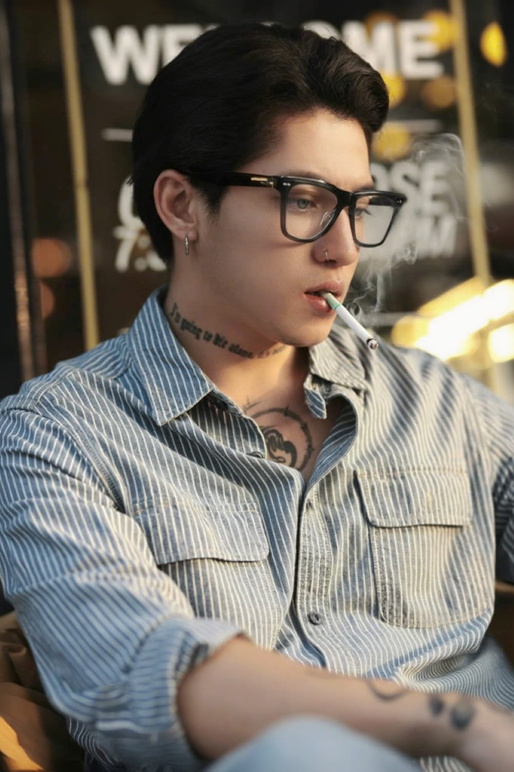 Person wearing Gentle Monster - GM Alto 01 and a striped shirt with tattoos, sitting indoors.