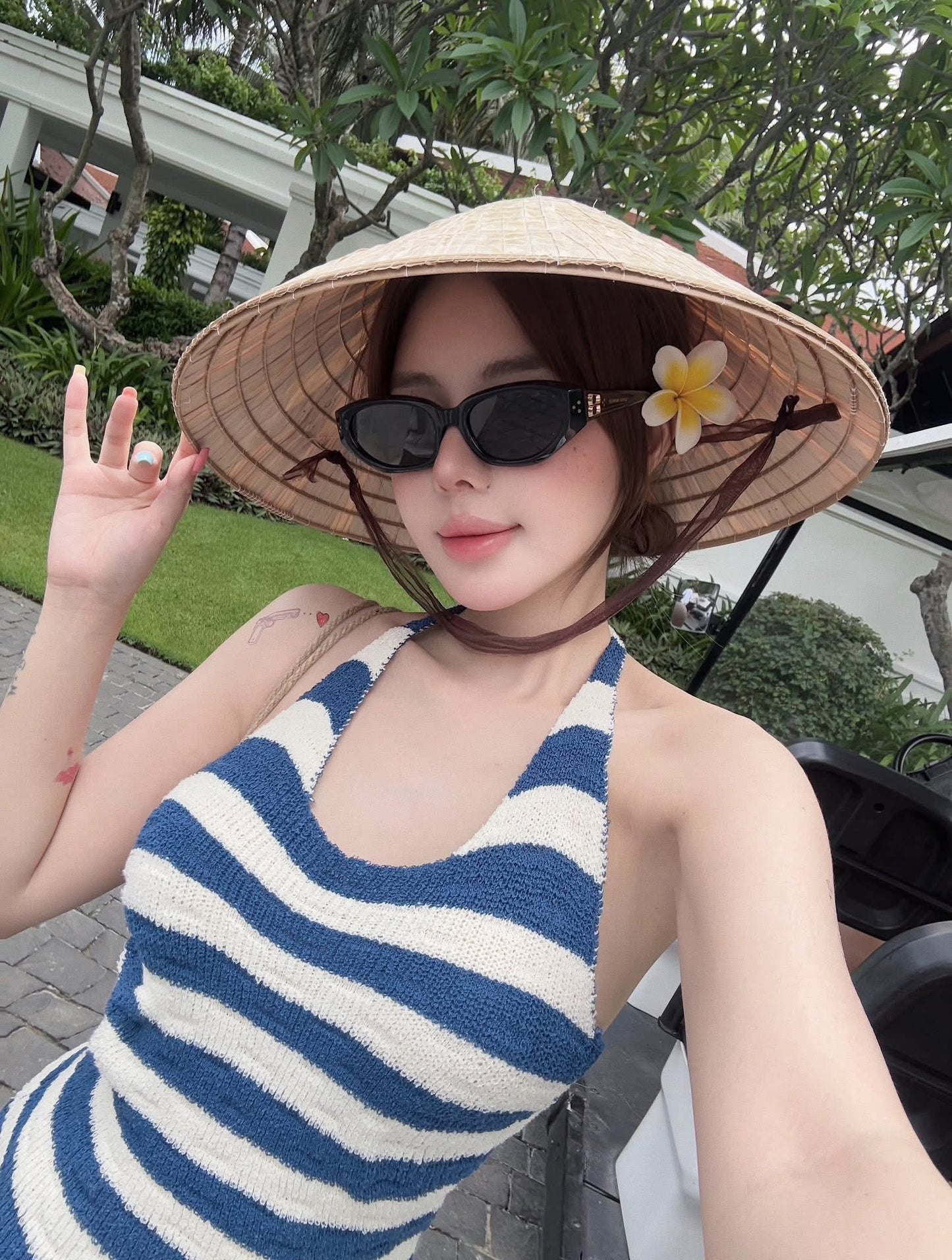 Woman wearing a straw hat with a flower, Gentle Monster (GM) Duns 01, and a blue and white striped dress outdoors.