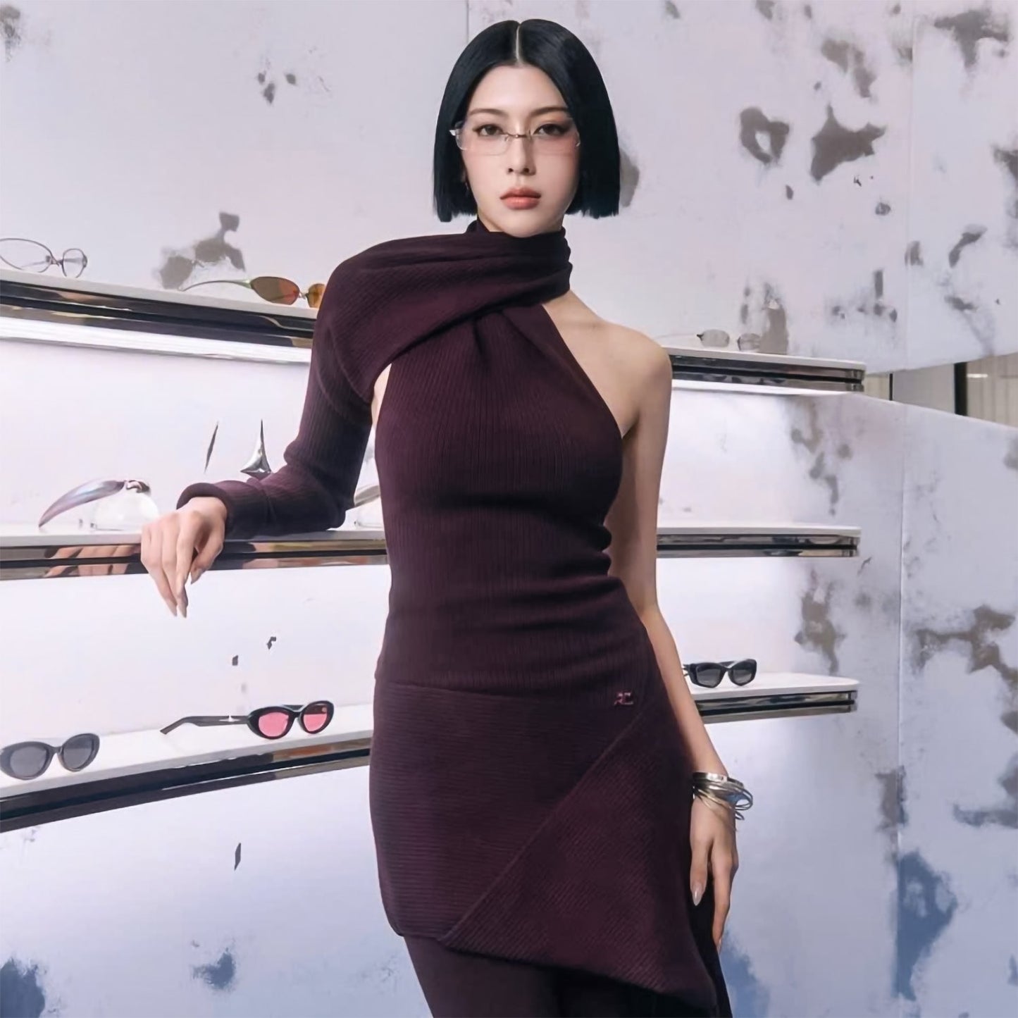 Woman wearing Gentle Monster Paranoyd 02(CM) Square Glasses in a burgundy dress standing in front of a display of sunglasses.