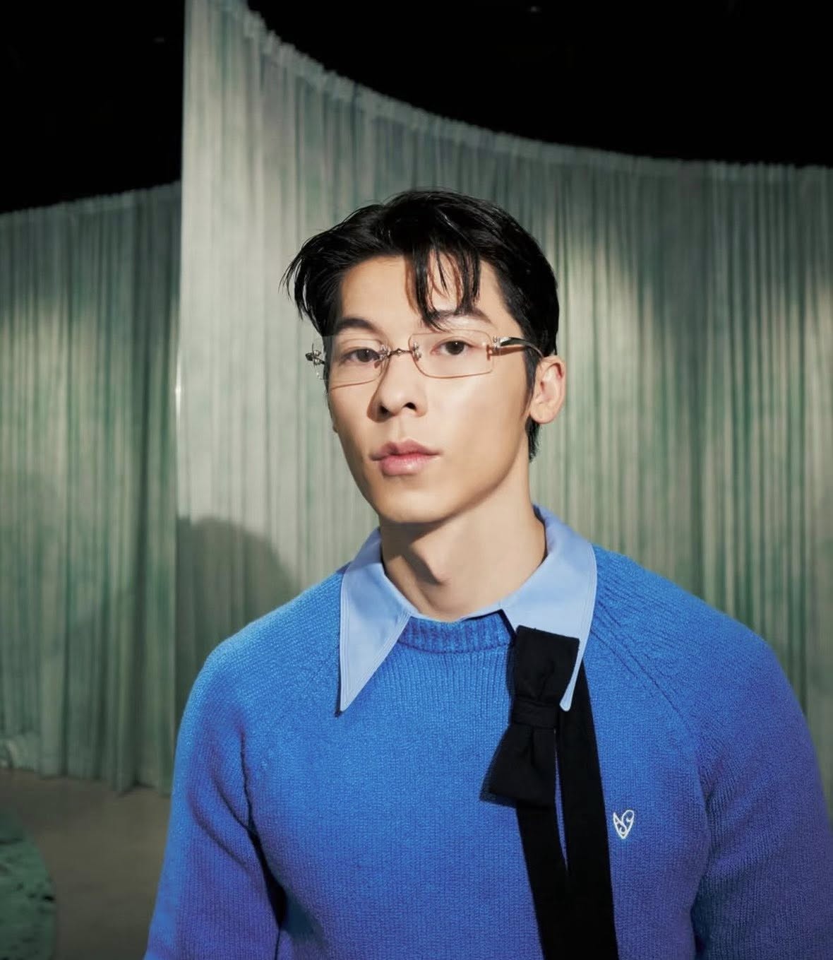 Greg Hsu / Greg Han wearing a Gentle Monster Paranoyd 02(CM) Square Glasses and blue sweater with a black tie against a green curtain background