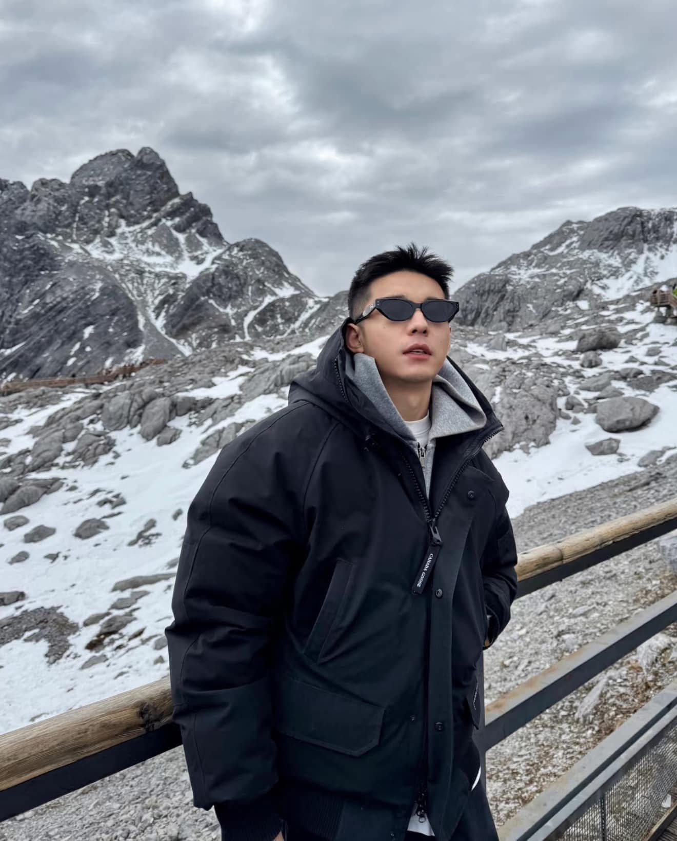 Person wearing Gentle Monster (GM) Rococo 01 and a black jacket standing in front of snow-covered mountains.