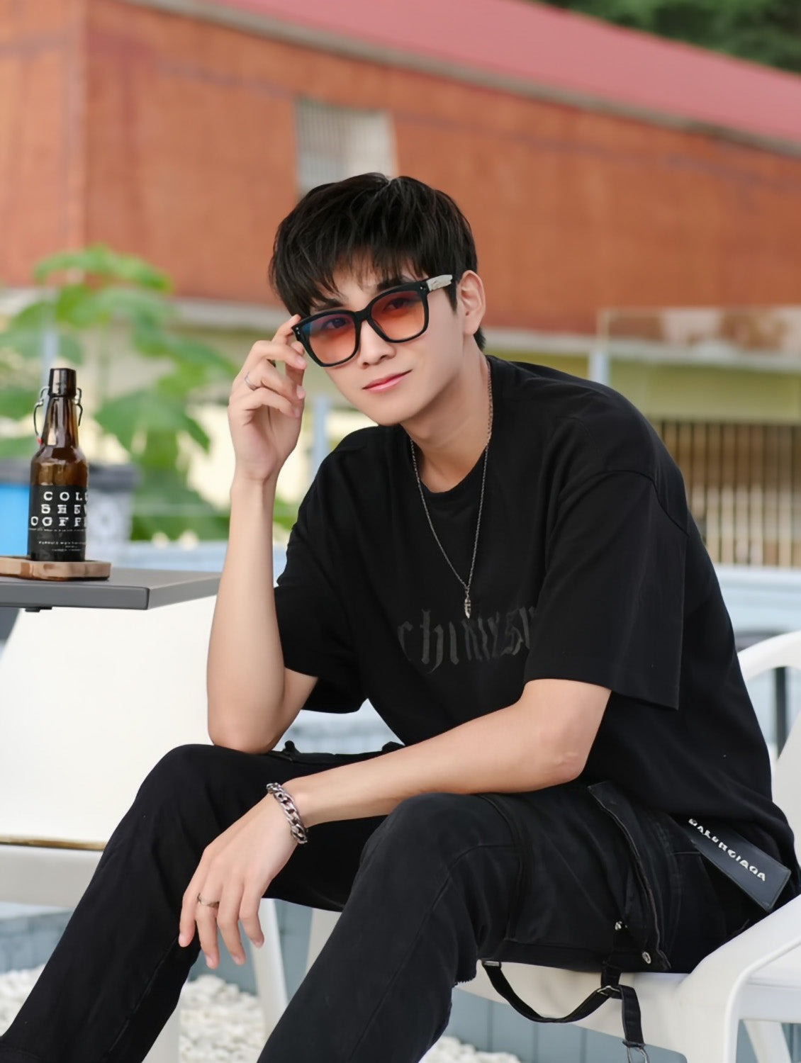 Person wearing Gentle Monster Una.C N 01(OR) Square Glasses sitting outdoors with a bottle of Cola Coffee in the background