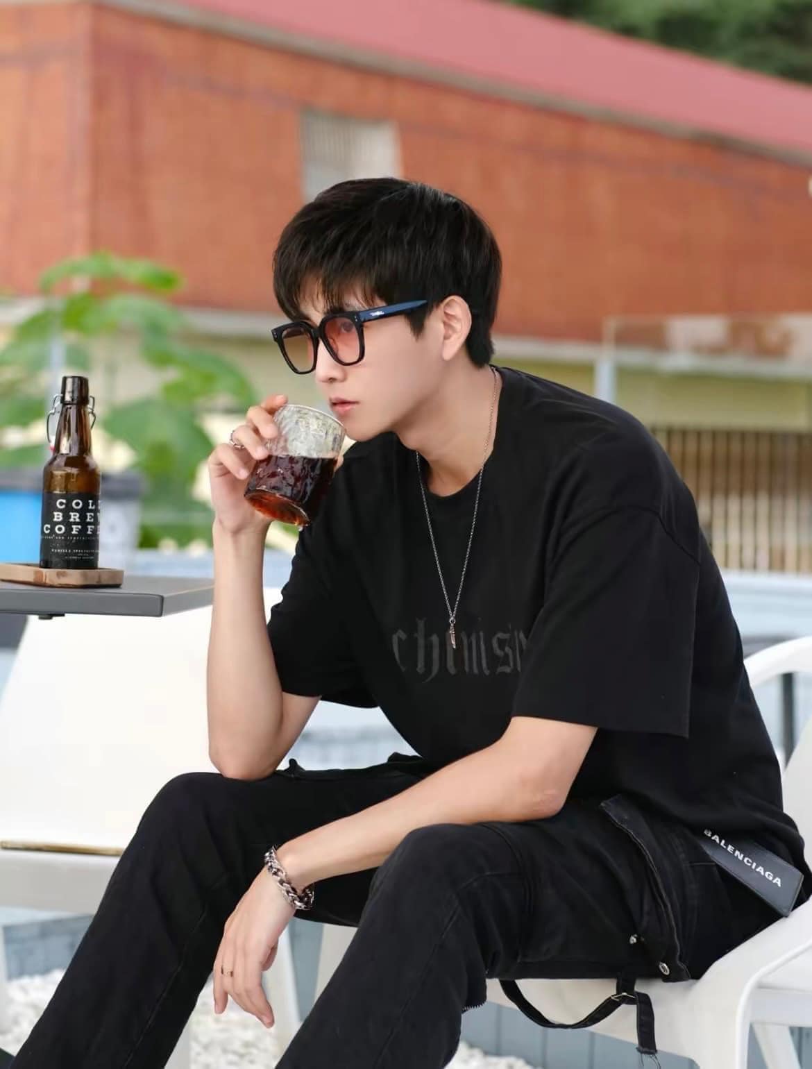 Person wearing Gentle Monster Una.C N 01(OR) Square Glasses in black outfit drinking from a glass outdoors