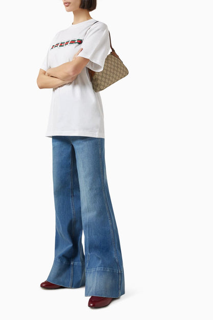 Person wearing a white t-shirt with text and blue jeans, holding a Gucci GG Emblem Small Shoulder Bag Beige 815218 FAEAA 9762 on a white background