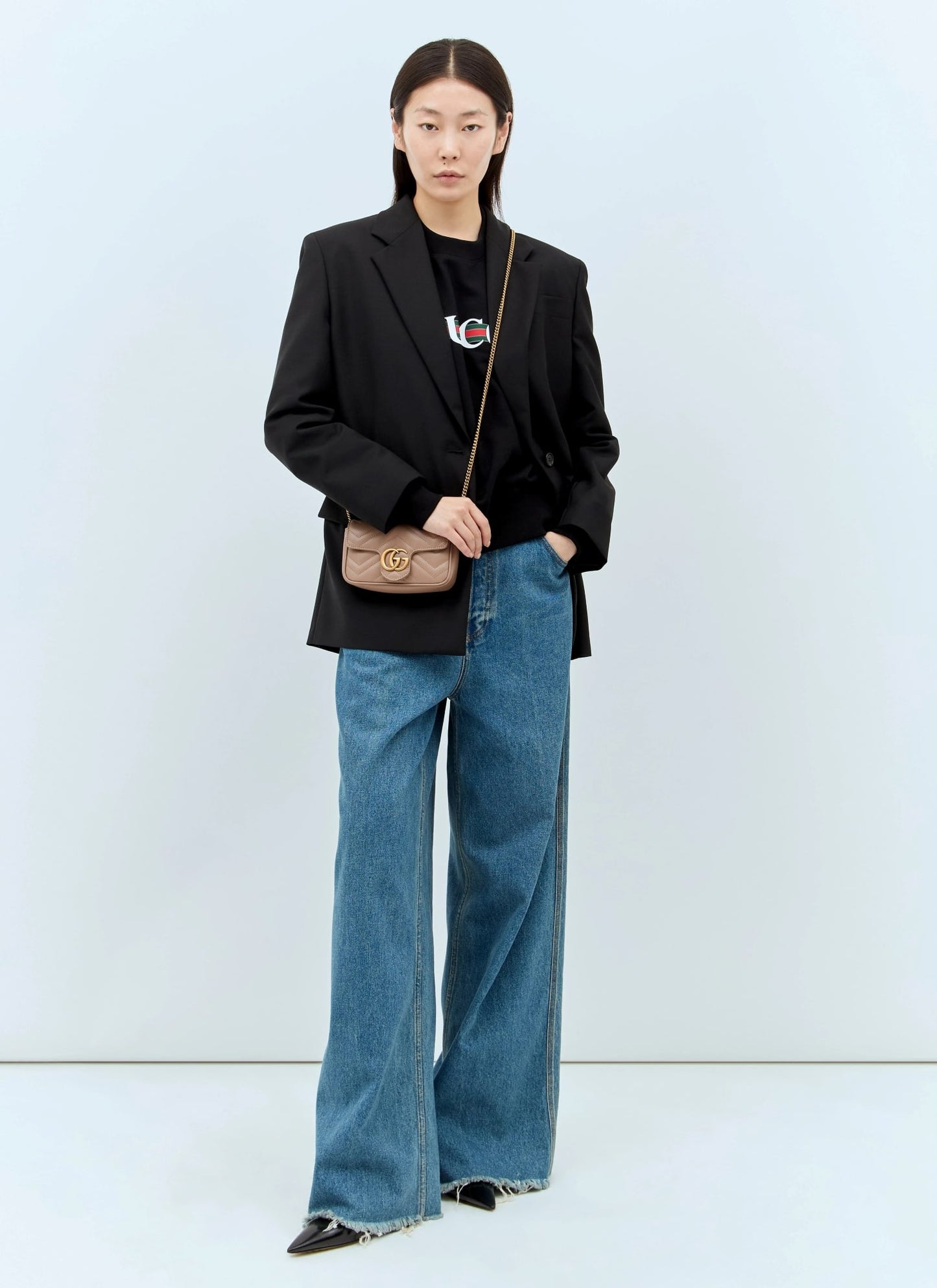 Person wearing a black blazer and blue jeans with a Gucci GG Marmont Matelassé Leather Mini Bag Dusty Pink 476433 DTDCT 5729 on a light blue background