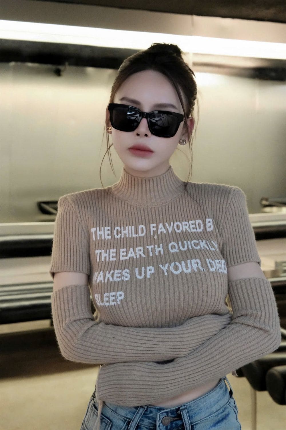 A woman wearing a beige ribbed turtleneck sweater with white text and cutout sleeves, paired with blue jeans and Gucci GG0001SN Black Rectangular Sunglasses, standing in an indoor setting.