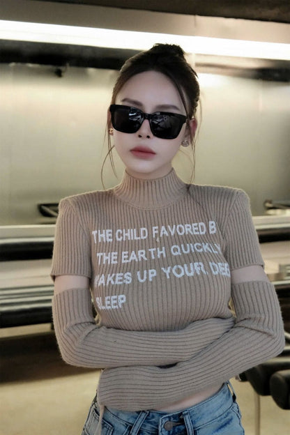 A woman wearing a beige ribbed turtleneck sweater with white text and cutout sleeves, paired with blue jeans and Gucci GG0001SN Black Rectangular Sunglasses, standing in an indoor setting.