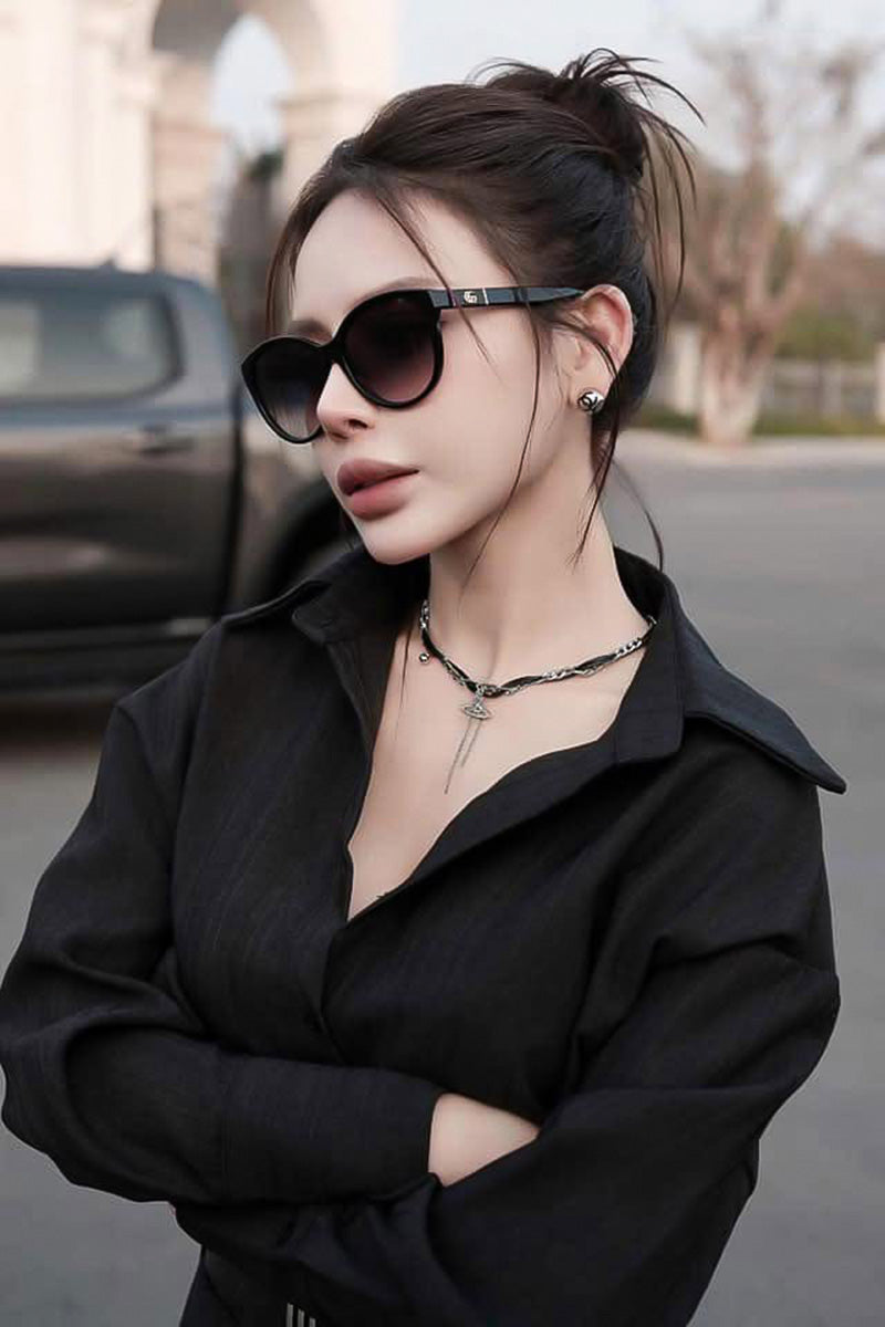 A woman wearing Gucci GG0636SK 001 Black Round Sunglasses, a black collared shirt, and a silver chain necklace, standing outdoors with her arms crossed.