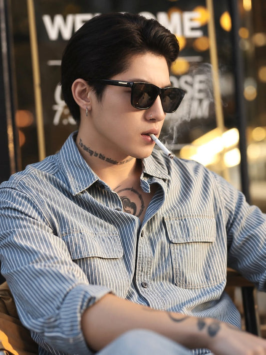 A man wearing a light blue and white vertically striped button-up shirt, Gucci GG0746S Black Square Sunglasses, and smoking a cigarette, set against a warm, blurred background with cafe signage.