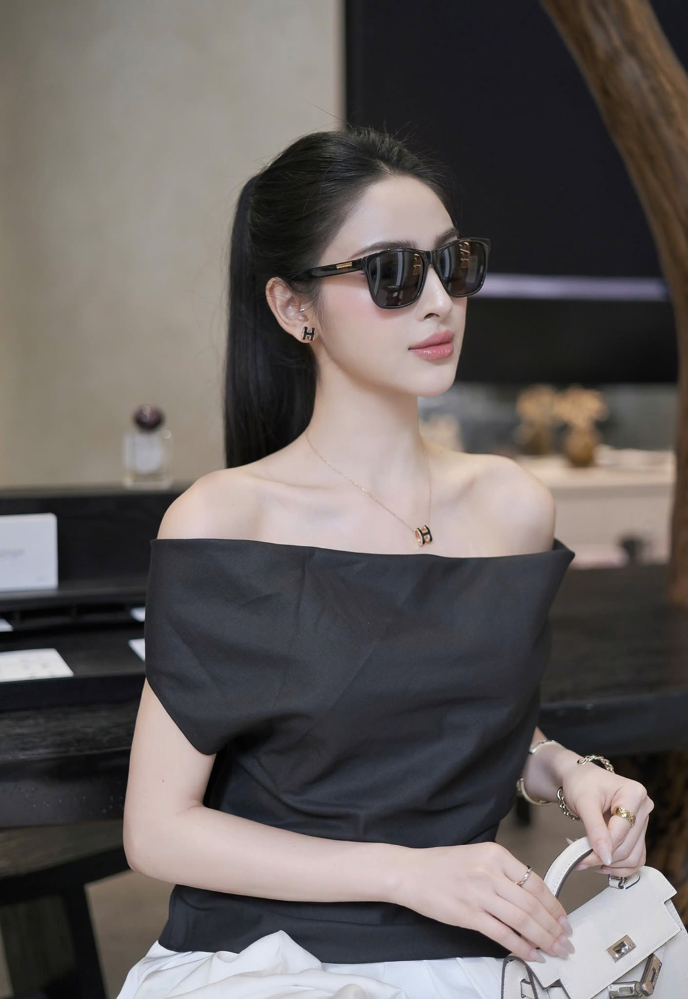 A woman wearing a black off-the-shoulder top, Gucci GG0746S Black Square Sunglasses, and a delicate gold necklace, holding a white handbag in an indoor setting.