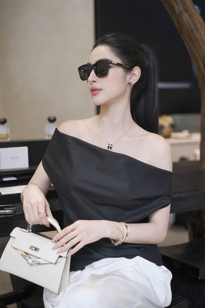 A woman wearing a black off-the-shoulder top and white bottoms, accessorized with Gucci GG0746S Black Square Sunglasses, a delicate necklace, and bracelets, holding a cream-colored handbag.