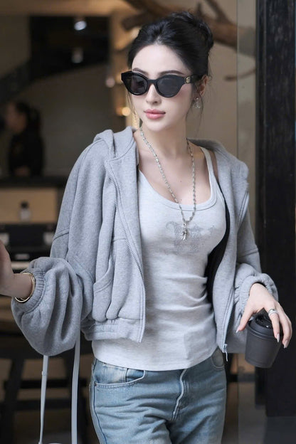 A woman wearing a light gray oversized zip-up hoodie over a white tank top and blue jeans, styled with Gucci GG0957S 002 Black Butterfly Rounded Sunglasses, a silver necklace, and holding a black travel tumbler in a modern indoor setting.