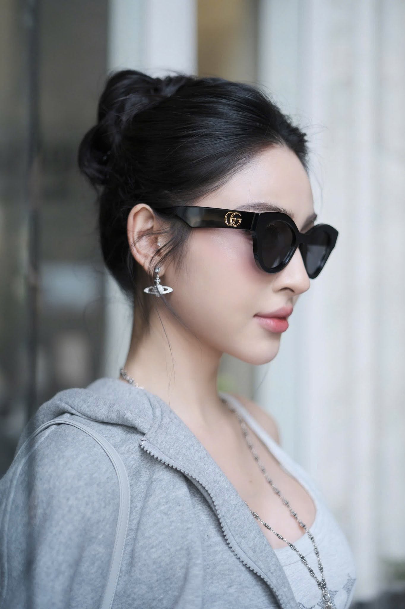 A woman in profile wearing Gucci GG0957S 002 Black Butterfly Rounded Sunglasses with gold double-G logo detailing on the temples, styled with a gray zip-up hoodie and silver jewelry.