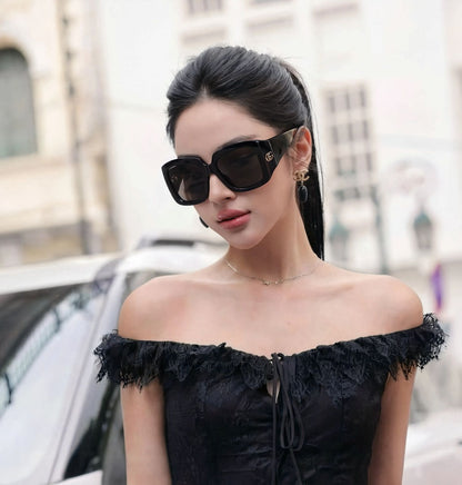 A woman wearing Gucci GG1402S 001 Black Square Oversized Sunglasses and a black off-the-shoulder lace top, photographed outdoors with a blurred city street background.