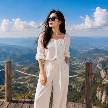 A woman wearing a Gucci GG1430SK 001 Black Squared Sunglasses in white long-sleeve blouse and matching white wide-leg trousers stands on a wooden viewing platform overlooking a vast mountain valley under a blue sky with clouds.