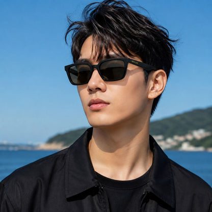 A man wearing Gucci GG1539S 001 Black Rectangular Sunglasses with dark lenses and a black shirt, photographed outdoors against a blue sky and sea background.