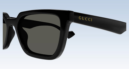 Gucci GG1539S 001 Black Rectangular Sunglasses with a glossy black frame and dark gray lenses, featuring the gold 'GUCCI' logo on the temple arm, set against a light blue gradient background.