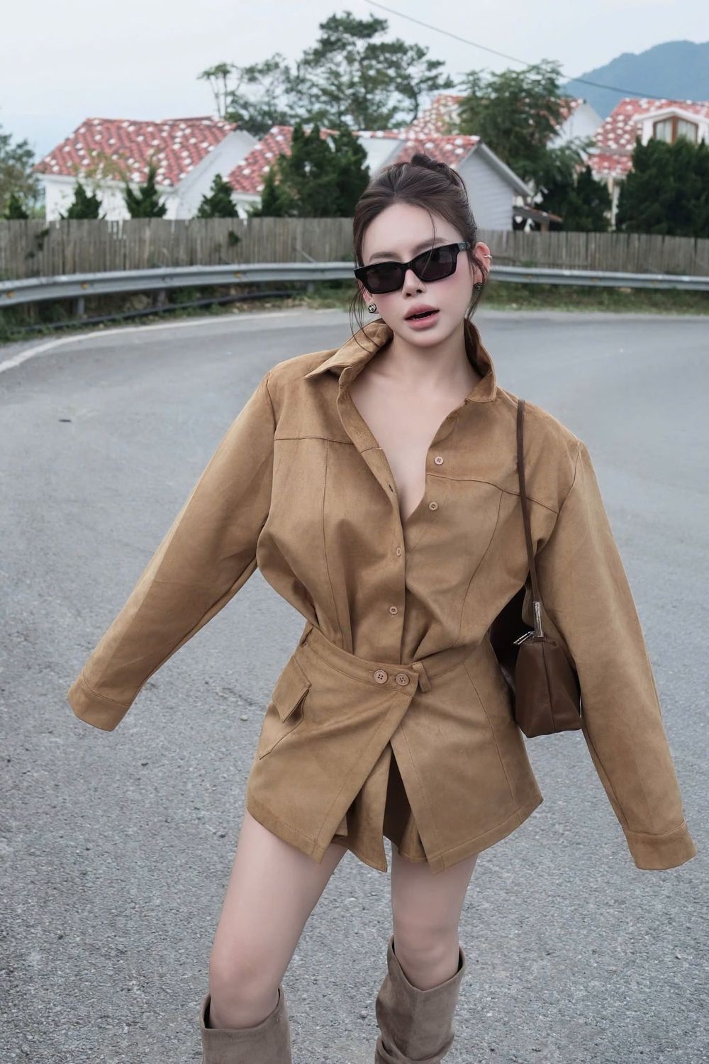 A woman wearing a Gucci GG1790S 001 Black Rectangular Sunglasses in tan-brown long-sleeve belted shirt dress and matching brown boots, standing on an outdoor road with houses and trees in the background.