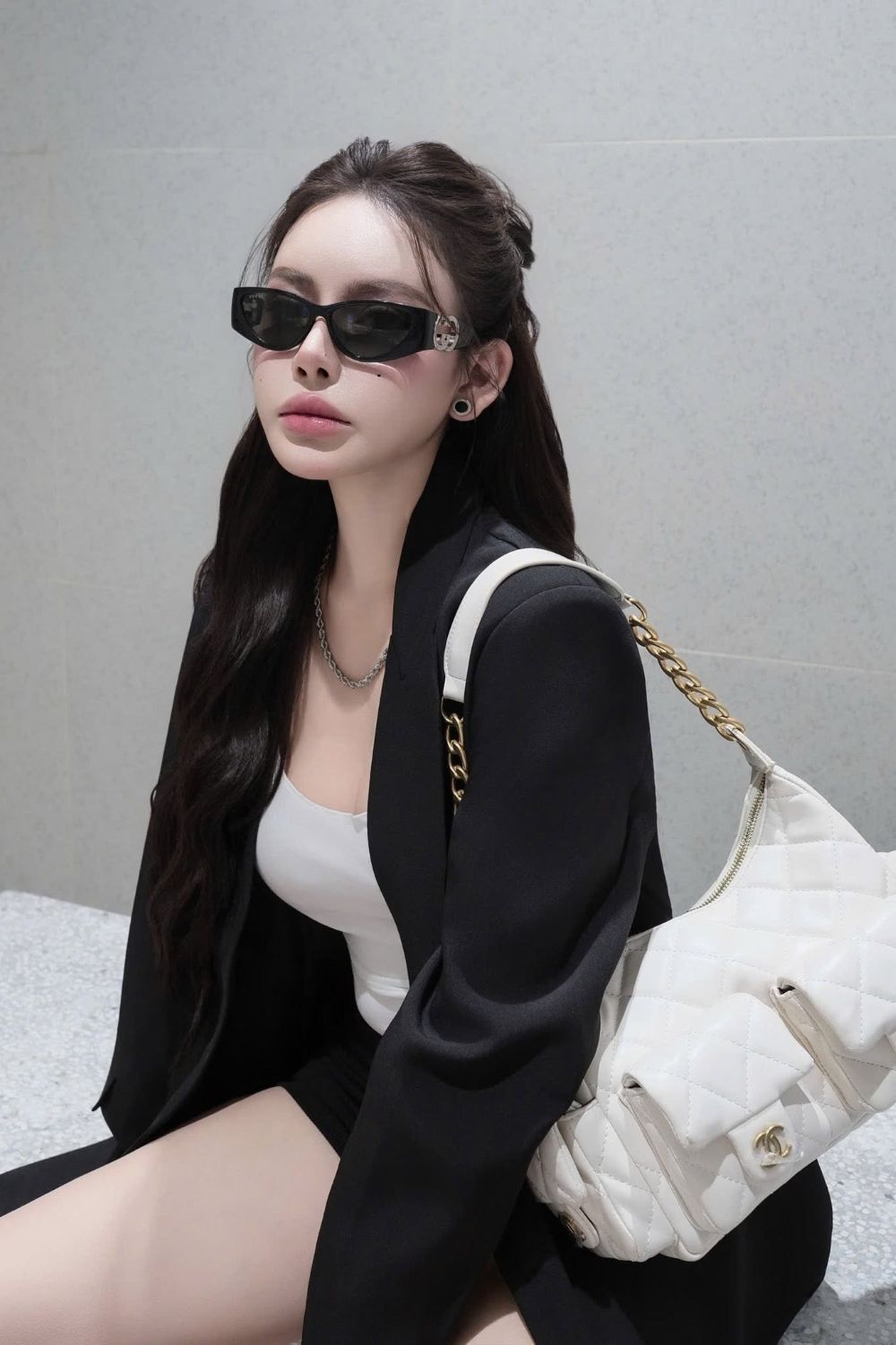 A woman wearing Gucci GG1824S 001 Black Cat-Eye Sunglasses, a black blazer over a white top, and holding a white quilted shoulder bag with a gold chain strap, posed against a gray tiled wall.