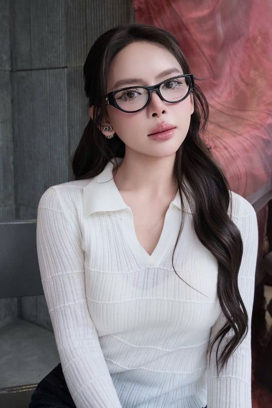 A woman with long dark wavy hair wearing Gucci GG1825O 001 Black Rounded Rectangular Eyeglasses and a white ribbed long-sleeve polo top, posed against a gray stone wall with a pink abstract backdrop.