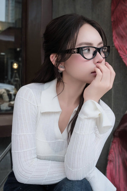 A woman wearing Gucci GG1825O 001 Black Rounded Rectangular Eyeglasses and a white ribbed long-sleeve collared top, posing in front of a dark window.