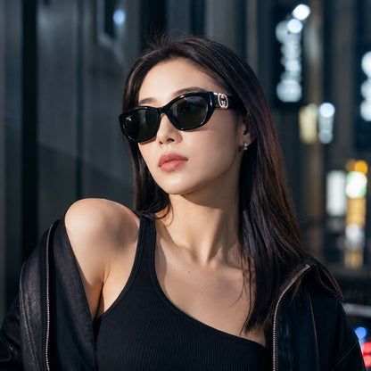 A woman wearing Gucci GG1826SK 001 Black Cat-Eye Sunglasses with a silver-tone GG logo on the temples, styled with a black ribbed top and an open black leather jacket against a blurred nighttime city street background.