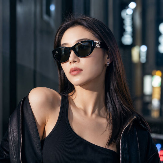 A woman wearing Gucci GG1826SK 001 Black Cat-Eye Sunglasses with a silver-tone GG logo on the temples, styled with a black ribbed top and an open black leather jacket against a blurred nighttime city street background.