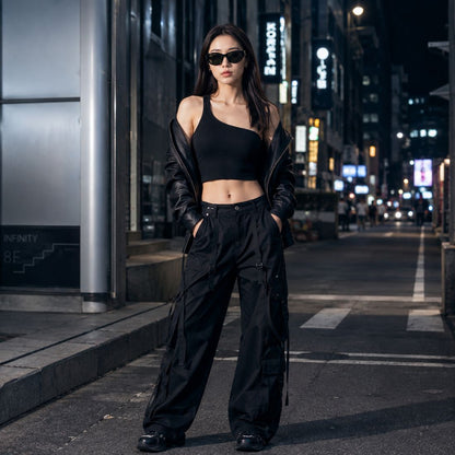 A woman stands on a city street at night wearing a Gucci GG1826SK 001 Black Cat-Eye Sunglasses in black asymmetrical one-shoulder top, black wide-leg cargo pants, a black leather jacket draped off her shoulders, and black sunglasses.