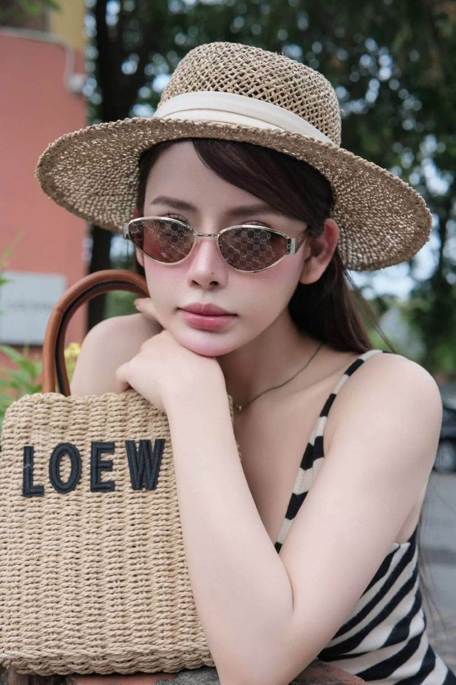 A woman wearing a straw hat, Gucci GG1853S 003 Gold Butterfly Sunglasses — Brown GG Lens, and a striped top holds a woven straw LOEWE handbag in an outdoor setting.