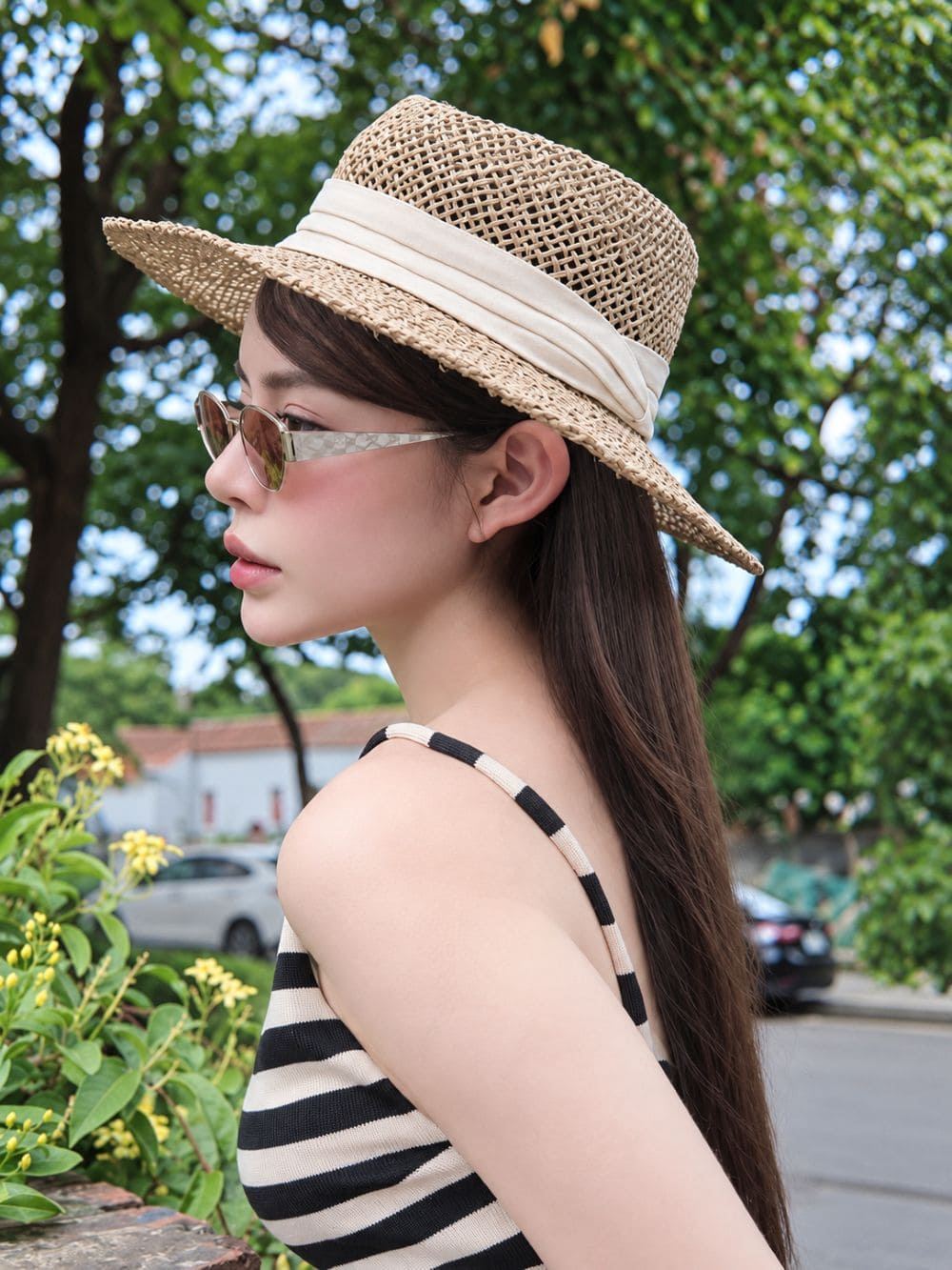 A woman in profile wearing a woven straw boater hat with a cream ribbon band, Gucci GG1853S 003 Gold Butterfly Sunglasses — Brown GG Lens, and a black-and-white striped top, photographed outdoors with trees and a street in the background.