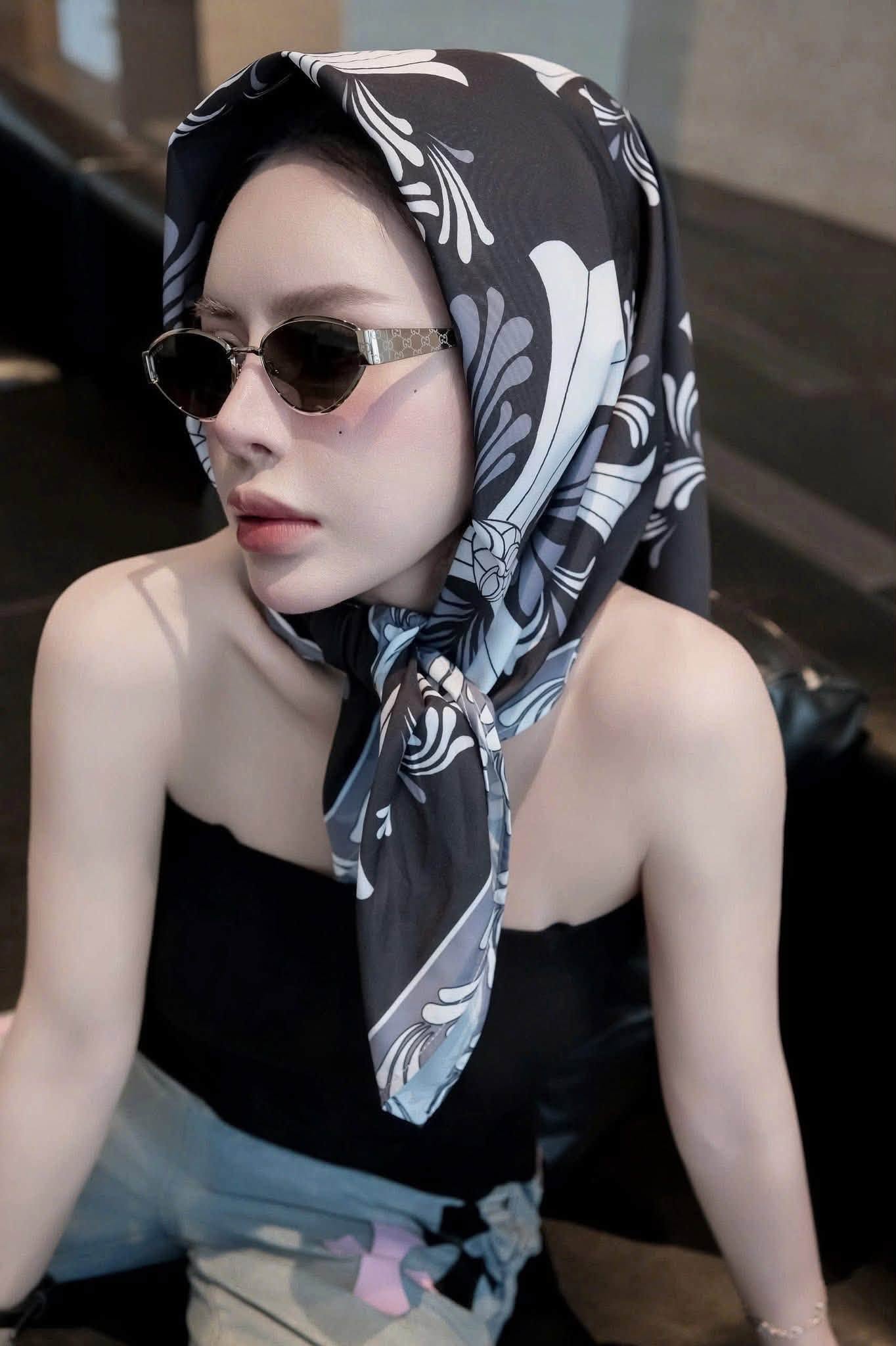 A woman wearing a black, white, and gray patterned headscarf and Gucci GG1853S 001 Gold Butterfly Sunglasses, posing in a dimly lit indoor setting.
