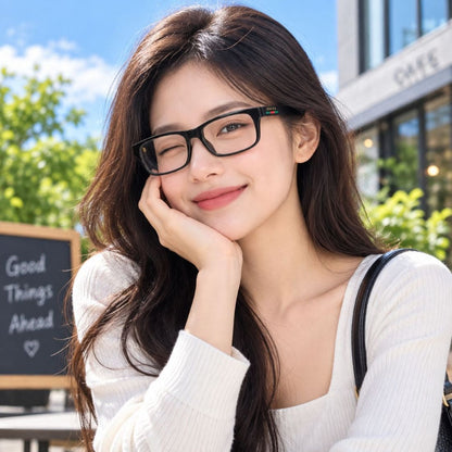 A woman with long dark brown hair wearing Gucci GG1858O Black Rectangular Eyeglasses and a white ribbed top, posing outdoors with a smile. The background shows a bright, sunny cafe or street setting with green foliage and a blue sky.