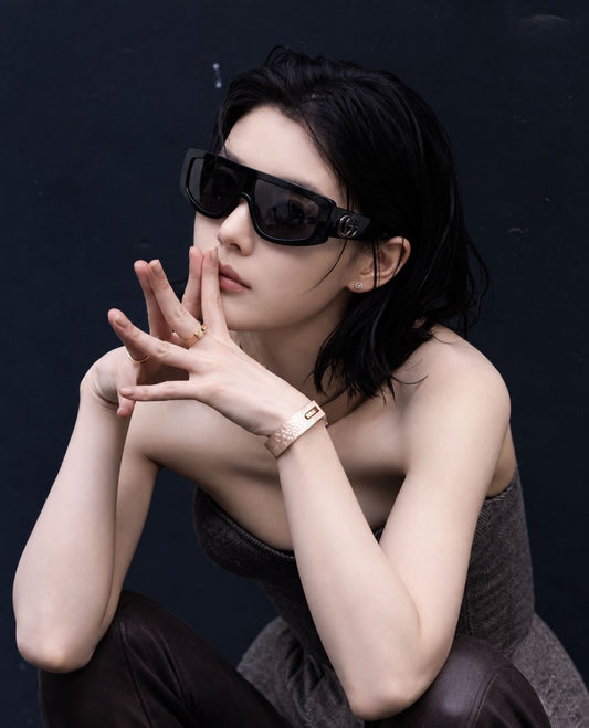 A woman wearing Gucci GG1950S 001 Black Shield Sunglasses, a gold bracelet, and a gold ring, posing against a dark textured background.