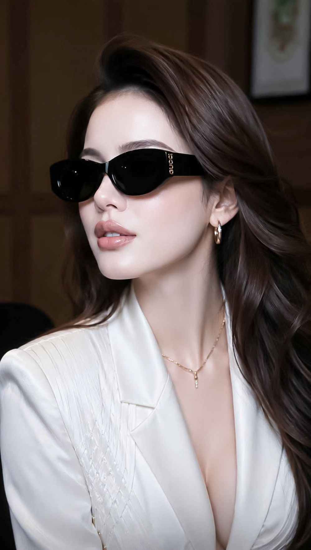 A woman with long brown hair wearing Gucci GG2044SA-001 Black Soft Rectangular Sunglasses with gold lettering on the temple, a white blazer, and gold jewelry, photographed in a warm indoor setting.