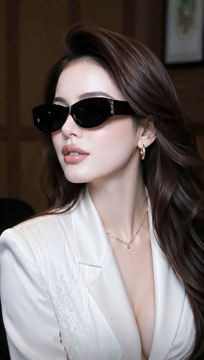 A woman with long brown hair wearing Gucci GG2044SA-001 Black Soft Rectangular Sunglasses with gold lettering on the temple, a white blazer, and gold jewelry, photographed in a warm indoor setting.