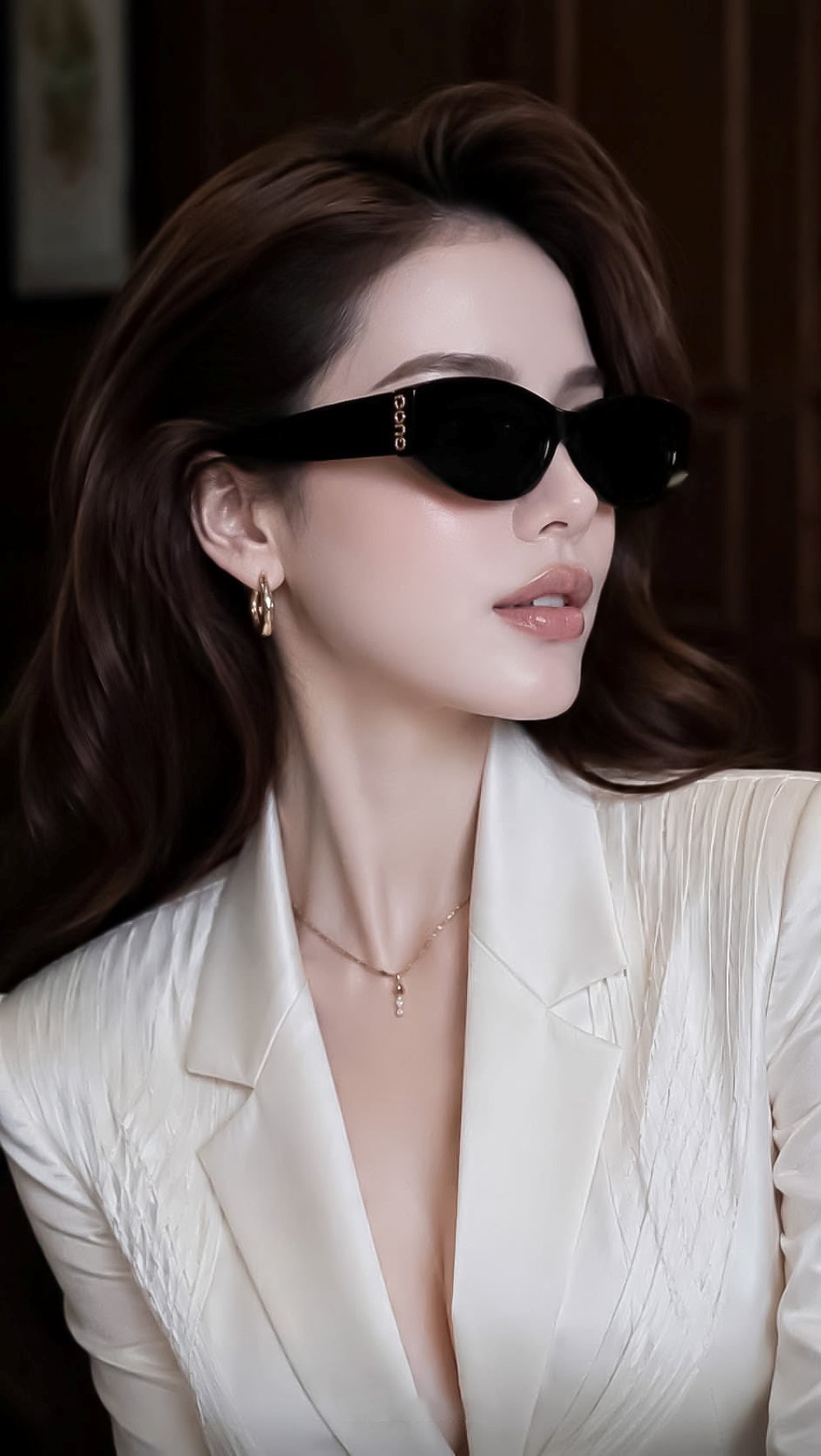 A woman with long brown hair wearing Gucci GG2044SA-001 Black Soft Rectangular Sunglasses with gold lettering on the temple, a cream-colored pleated blazer, and gold jewelry, set against a dark brown background.