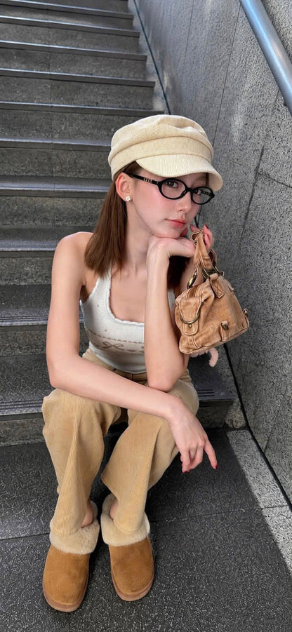 A woman sits on outdoor concrete stairs wearing a beige cap, Gucci GG2153O Round Eyeglasses, a white ribbed tank top, beige corduroy pants, tan suede boots, and holding a small brown leather shoulder bag.