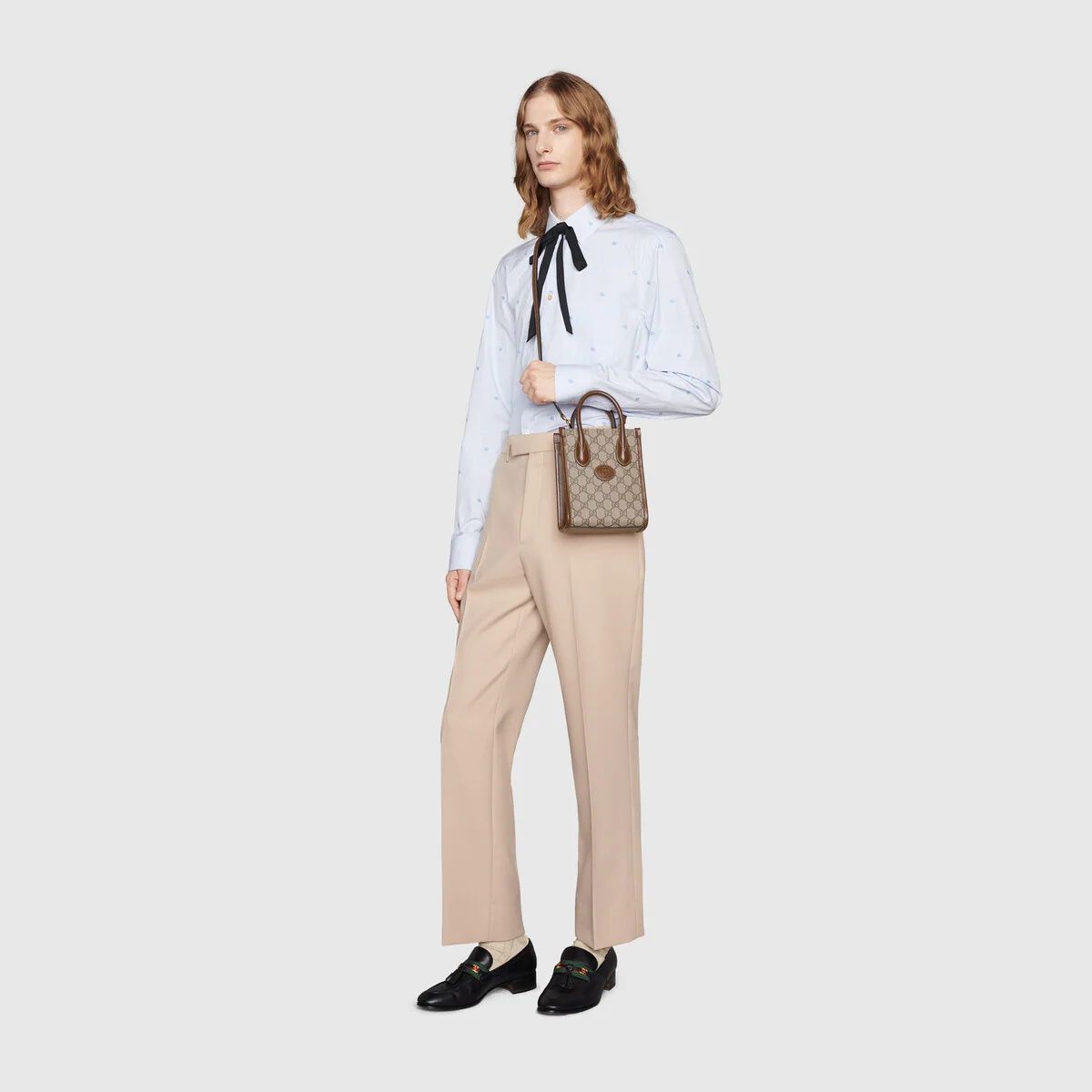 Woman wearing a white blouse with a black ribbon, beige pants, and holding a Gucci Interlocking G Mini Tote Bag 671623 92TCG 8563 on a light gray background