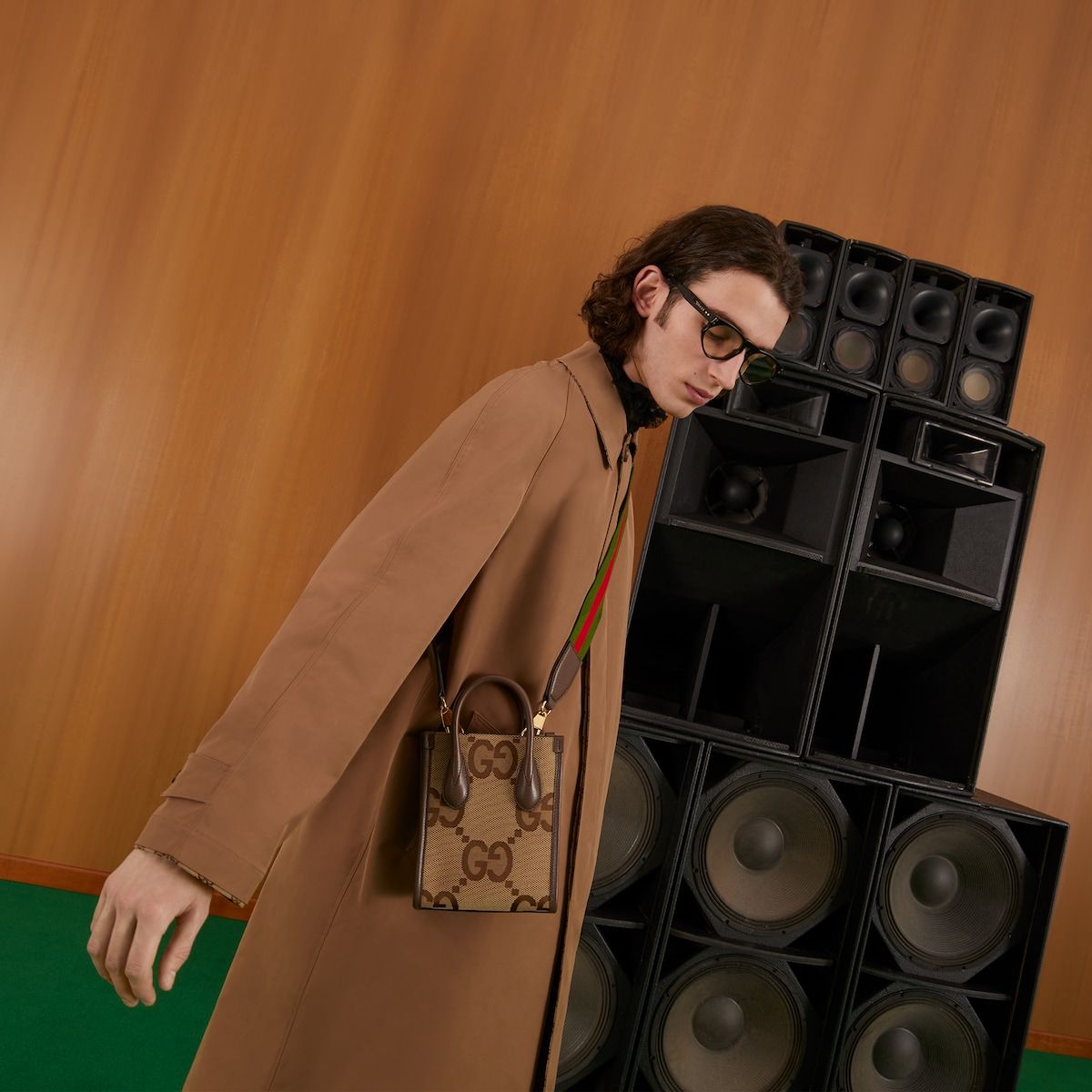 Person in a long coat with a Gucci Jumbo GG Mini Tote Bag Camel Ebony 699406 UKMDG 2570 standing next to a stack of speakers against a wooden wall.