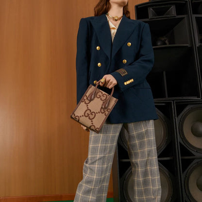 Person wearing a navy blue blazer with gold buttons, holding Gucci Jumbo GG Mini Tote Bag Camel Ebony 699406 UKMDG 2570, standing against a wooden wall with speakers.