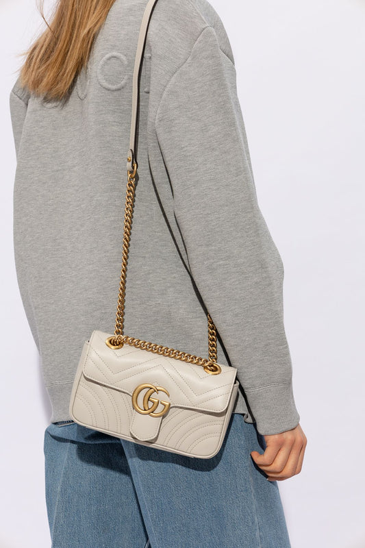Person wearing a gray sweater and blue jeans with a Gucci GG Marmont Small Shoulder Bag Light Grey 446744 AABZC 1712 featuring a gold chain strap on a white background